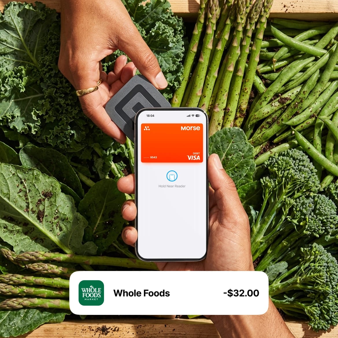 Hand tapping a phone with the Morse Card in Apple Pay against a tray of fresh greens, with a -$32 Whole Foods transaction
