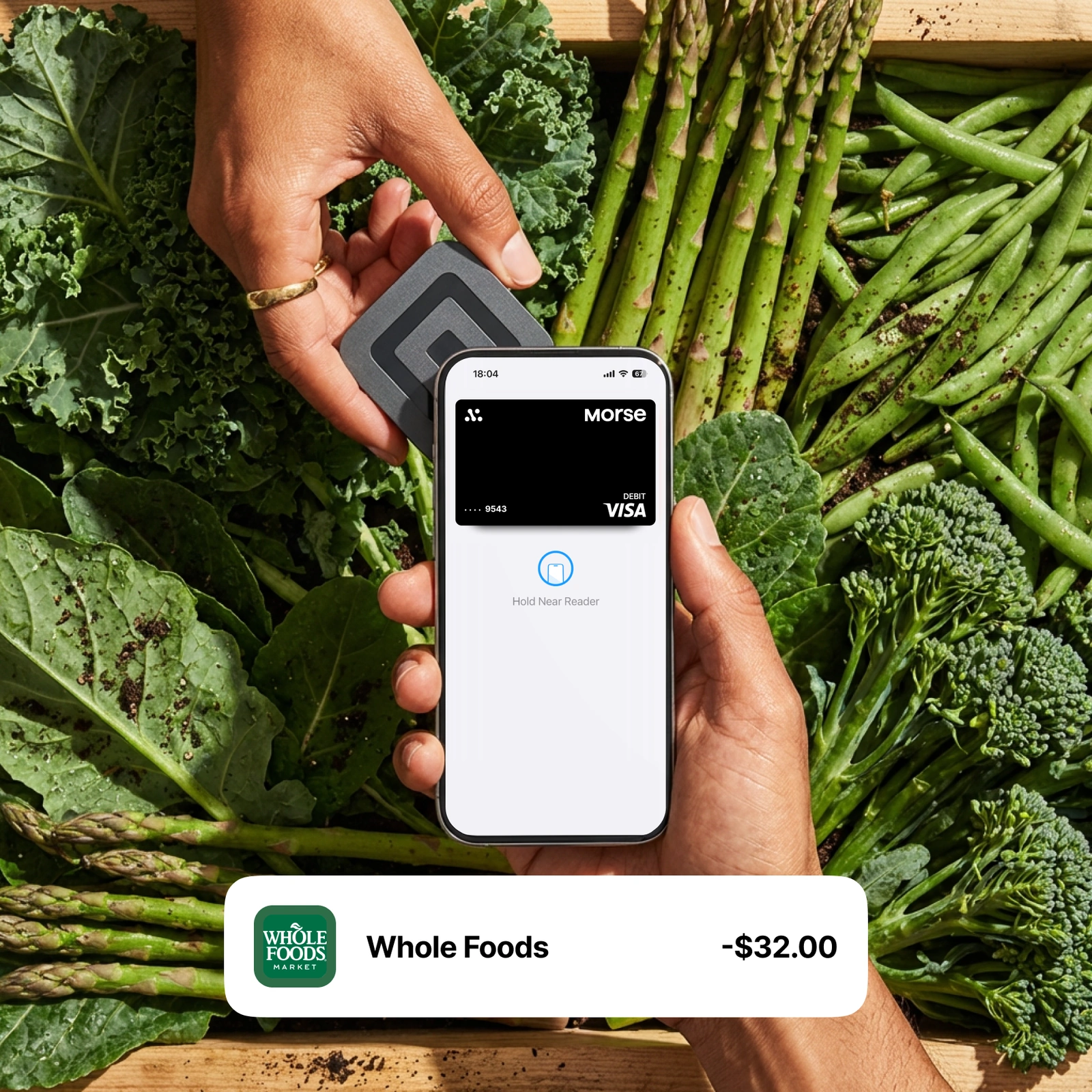 Hand tapping a phone with the Morse Card in Apple Pay against a tray of fresh greens, with a -$32 Whole Foods transaction